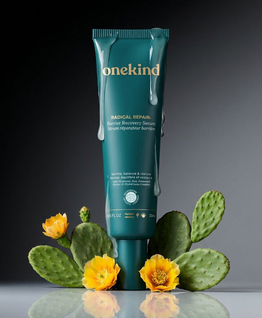A teal squeeze tube of Onekind Radical Repair Biotic Recovery Gelts centered on a reflective surface, surrounded by green cactus pads and bright yellow flowers against a dark gray to black gradient background.